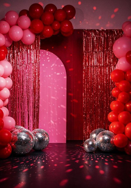Elegant red and pink balloon arch with shimmering foil curtains and disco balls for a stylish party backdrop.