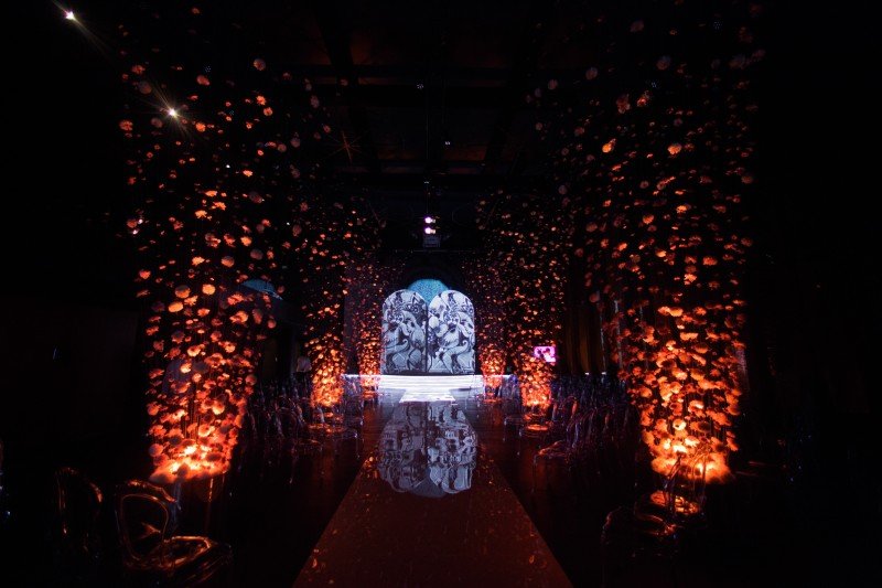 Dark, dramatic event venue with walls covered in glowing orange floral decorations, leading to a reflective aisle and an illuminated ornate backdrop.